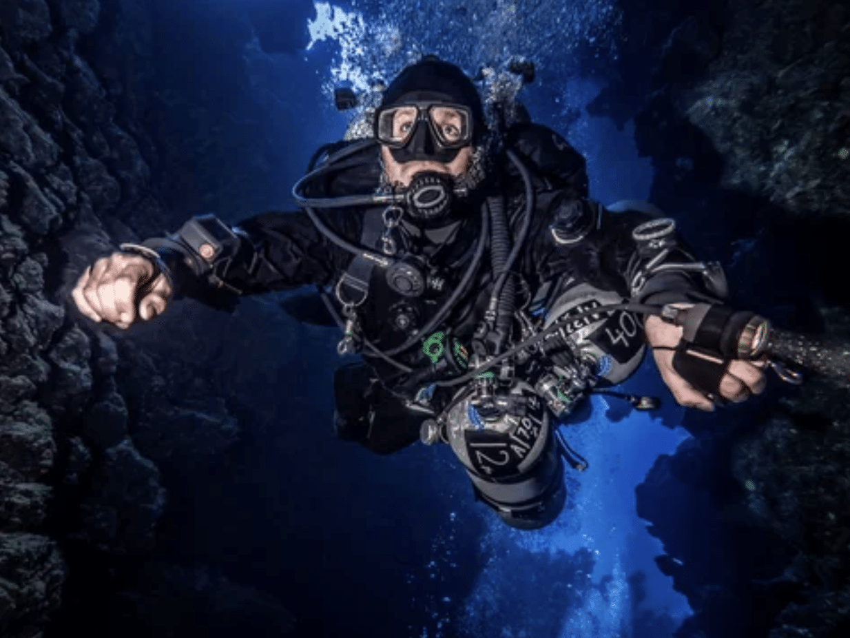 A scuba diver in full kit swims underwater between rocky walls, surrounded by bubbles, with rays of sunlight illuminating the blue water.