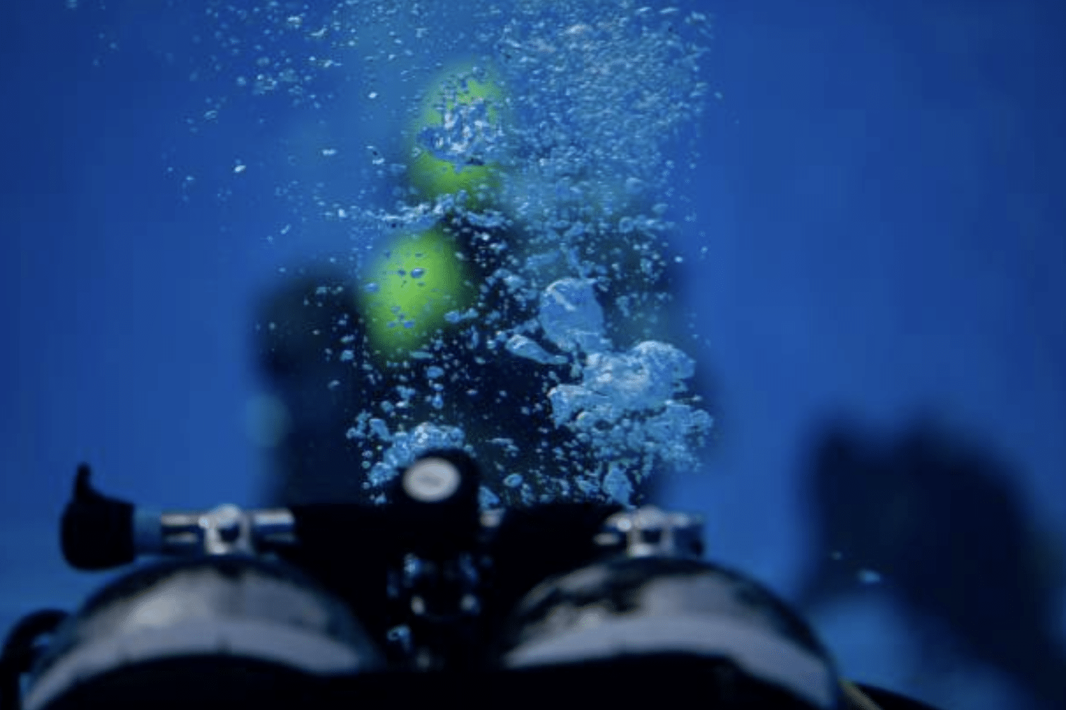 Screenshot 2026-04-19 at 194138 - N9BO℠ | Global Underwater Services Ltd A scuba diver is underwater, with air bubbles rising towards the surface. The image shows scuba cylinders in the foreground and the diver blurred in the background, surrounded by blue water.
