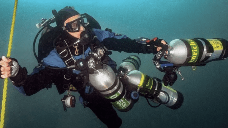 A scuba diver wearing a helmet and drysuit holds onto a yellow rope underwater, carrying multiple large cylinders labelled “enriched air” and technical diving equipment.