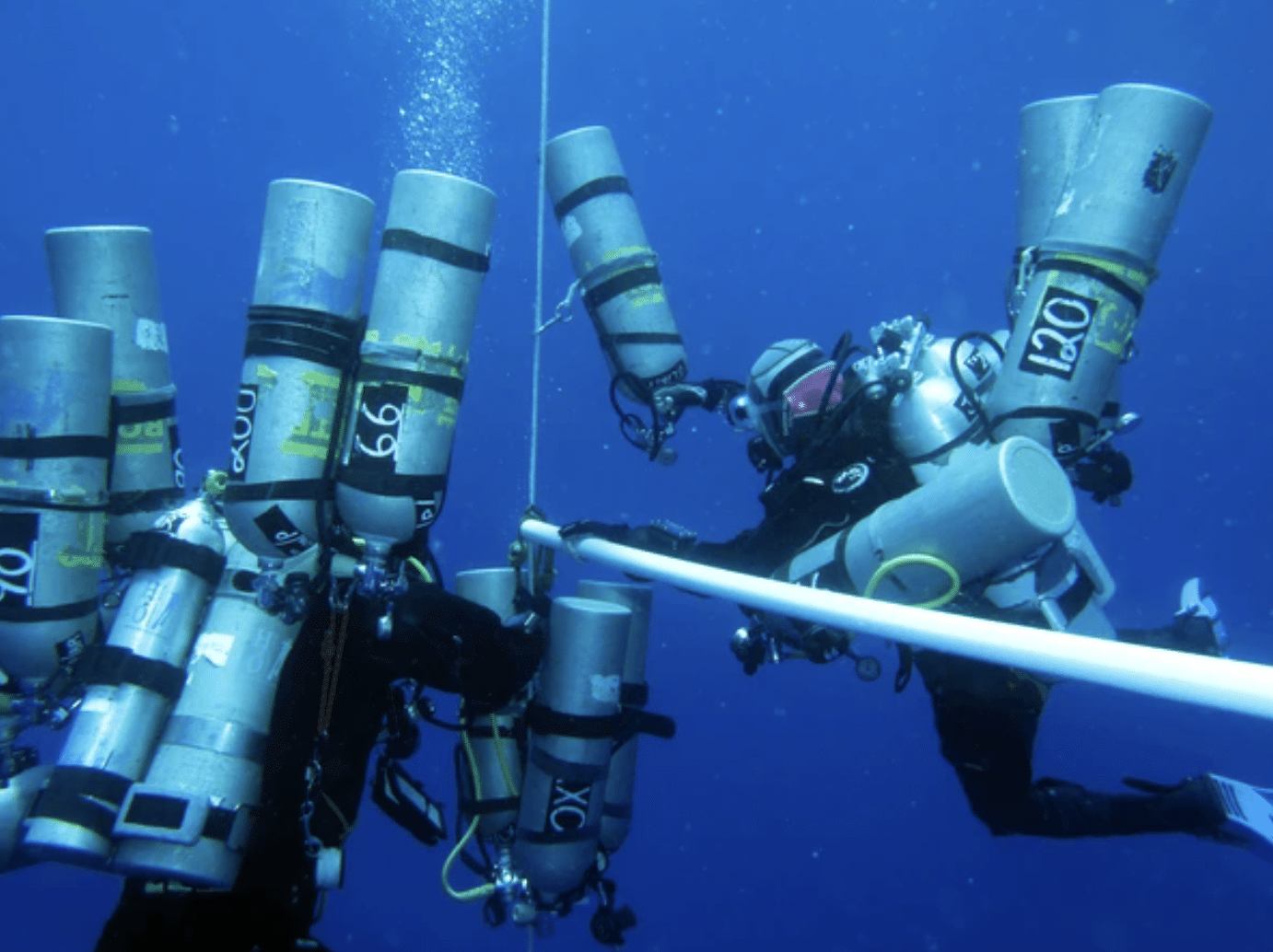 Screenshot 2026-04-19 at 204157 - N9BO℠ | Global Underwater Services Ltd Two scuba divers in full kit, carrying multiple large oxygen cylinders, are underwater, holding onto a white guide rope in deep blue water.
