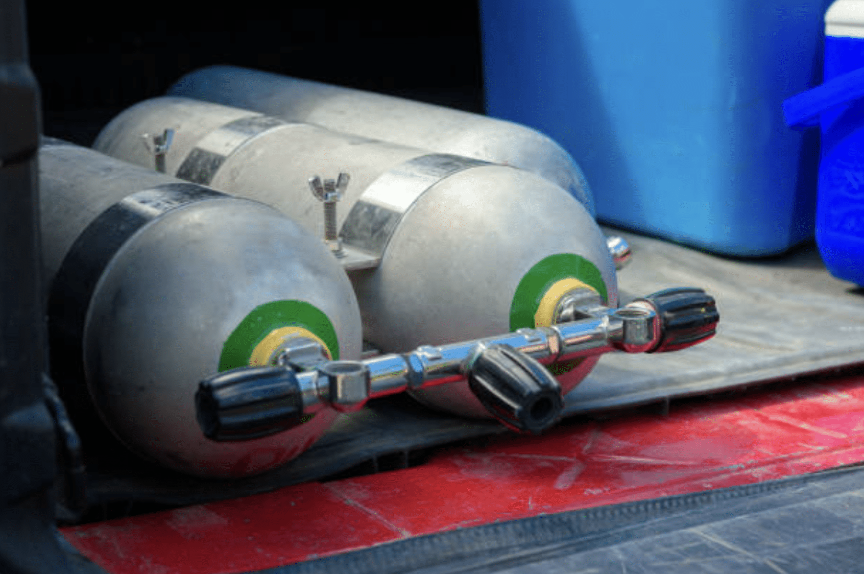 Two scuba diving air cylinders with green and yellow markings are lying on their sides next to blue cool boxes inside a vehicle, resting on a red and black surface.