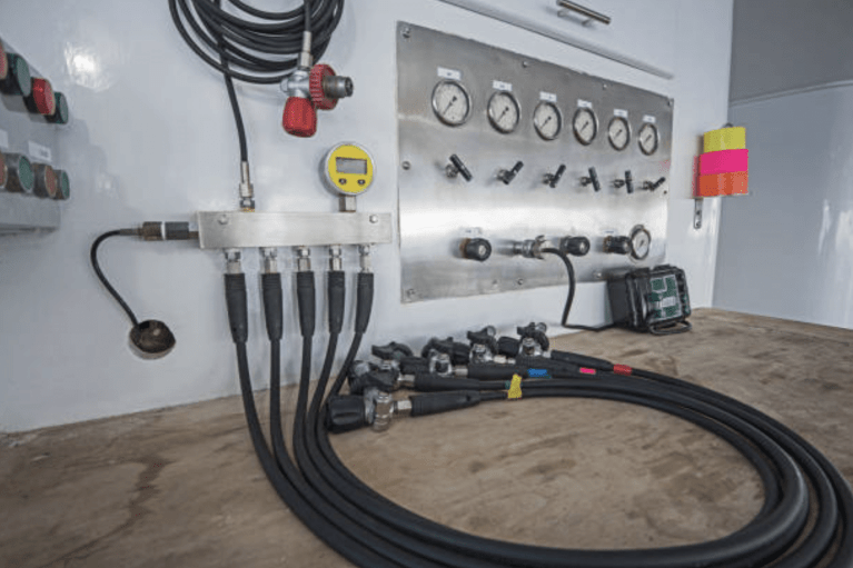 A control panel with gauges, switches, and multiple connected cables is mounted on a wall; brightly coloured markers and coiled cables are visible on a wooden surface in front of the panel.