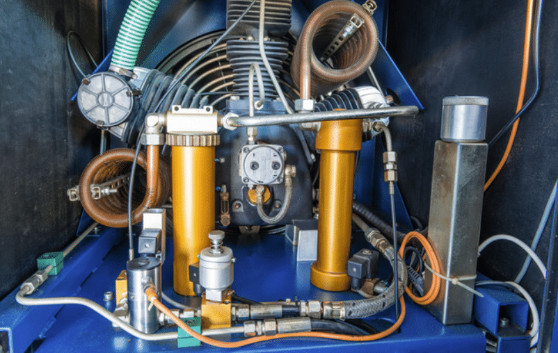 Screenshot 2026-04-24 103308 - N9BO℠ | Global Underwater Services Ltd Industrial hydraulic/pneumatic machinery with orange vertical cylinders, hoses, coils and metal components on a blue base, likely part of a compressor or pump system.