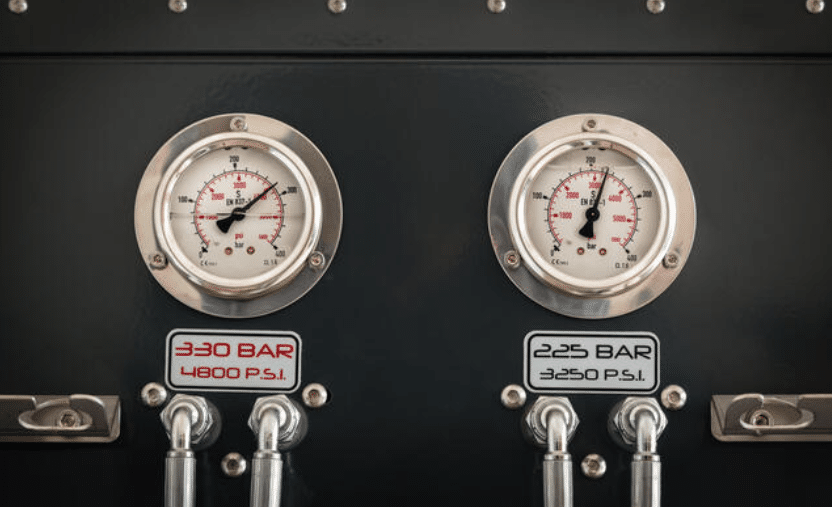 Two pressure gauges mounted on a panel display readings in bar and psi units. The left gauge shows 330 bar (4,800 psi), and the right gauge shows 225 bar (3,250 psi). Metal pipes are connected below each gauge.
