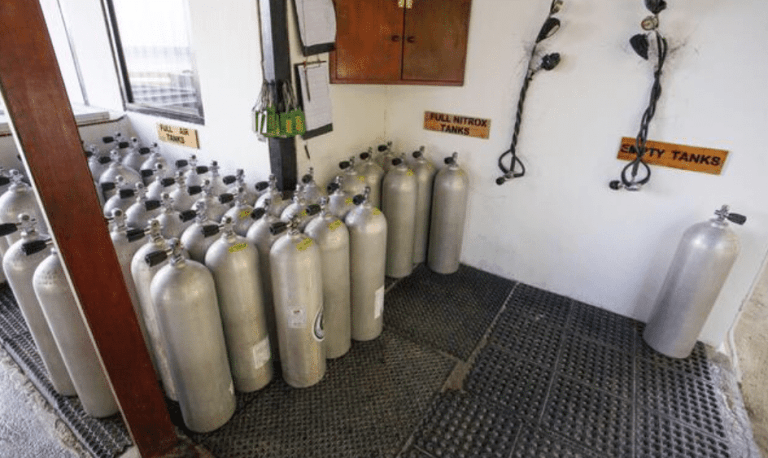 Rows of metal scuba cylinders stand upright on a rubber mat, with signs indicating sections for “Full Air,” “Full Nitrox,” and “Empty Cylinders” in a well-lit storage area. Some hoses hang on the wall.