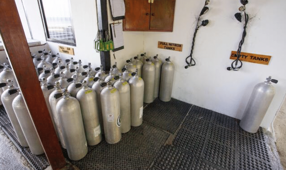 Screenshot 2026-04-24 103334 - N9BO℠ | Global Underwater Services Ltd Rows of metal scuba cylinders stand upright on a rubber mat, with signs indicating sections for “Full Air,” “Full Nitrox,” and “Empty Cylinders” in a well-lit storage area. Some hoses hang on the wall.