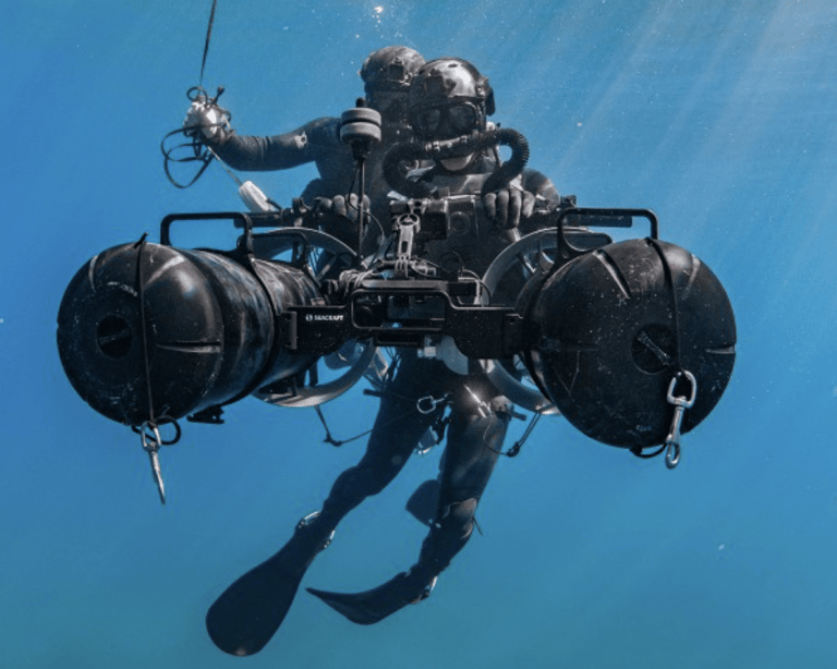 Two scuba divers in black gear use an underwater propulsion device in clear blue water, surrounded by sunlight streaming from the surface above.