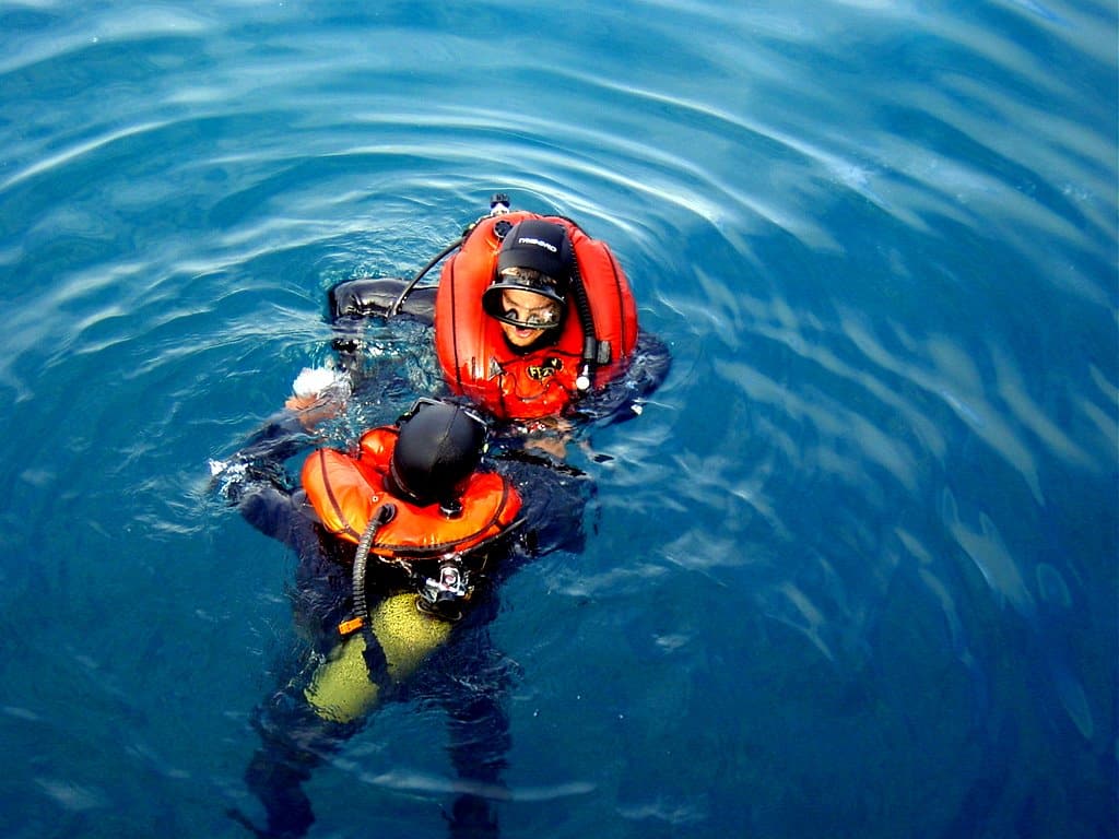 Scuba_divers_with_inflated_Fenzy-ABJL - N9BO℠ | Global Underwater Services Ltd Two people wearing diving gear and orange life jackets float in deep blue water. One appears to be assisting the other, who is on their back facing upward. The water around them is calm with gentle ripples.