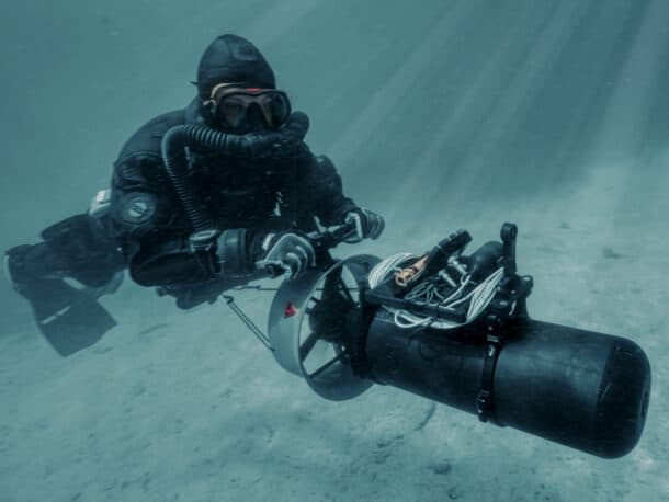 A scuba diver in a black wetsuit uses an underwater propulsion device to move along the sandy sea floor, surrounded by filtered sunlight.