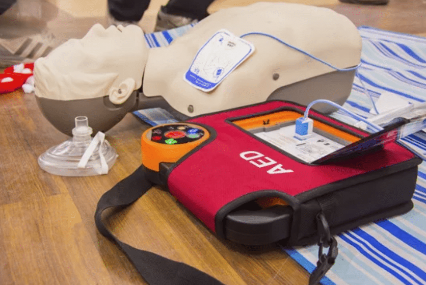 A CPR training manikin lies on a mat next to an automated external defibrillator (AED) and a clear plastic resuscitation mask, used for practising emergency response techniques.