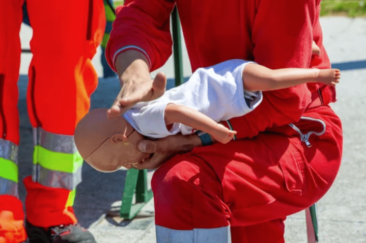 A person in red emergency uniform demonstrates infant first aid on a baby mannequin, holding it face down and patting its back, likely showing a choking rescue technique.