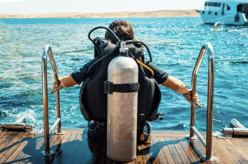 Back view of a scuba diver in full gear sitting on a boat ladder, ready to enter the clear blue water.