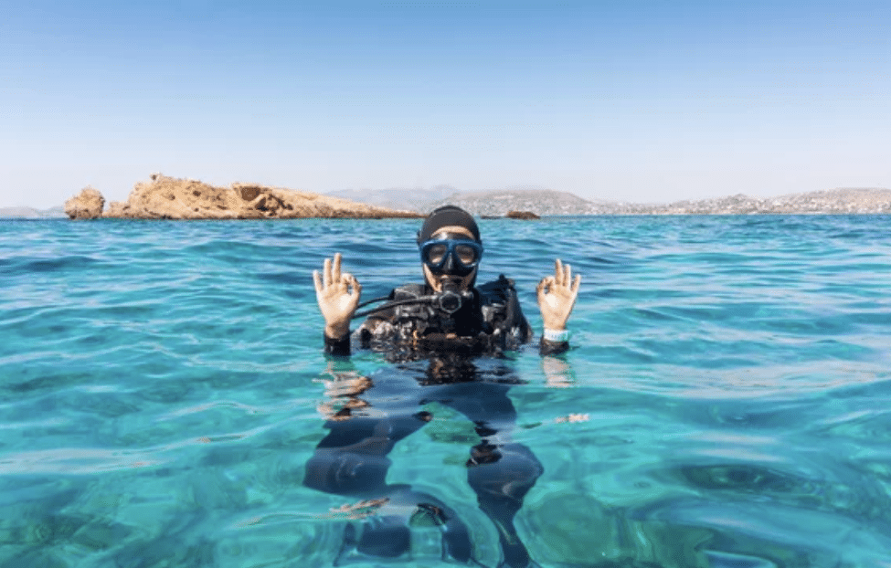 A scuba diver wearing a wetsuit, mask, and snorkel gives an “OK” hand signal while floating on the surface of bright blue water, with a rocky island and distant shoreline visible in the background.