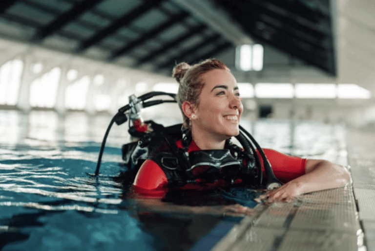 A smiling person in a red wetsuit and scuba gear is in an indoor swimming pool, holding onto the pool edge, with light streaming through large windows in the background.