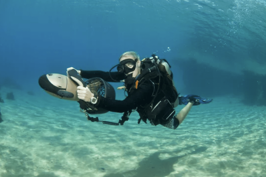 Screenshot 2026-05-18 154528 - N9BO℠ | Global Underwater Services Ltd A scuba diver wearing a black wetsuit, mask, and fins uses an underwater scooter to glide above the sandy sea floor, with clear blue water and faint outlines of rocks or structures in the background.