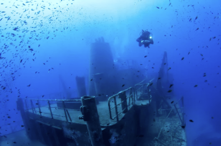 Screenshot 2026-05-18 154658 - N9BO℠ | Global Underwater Services Ltd A scuba diver explores a sunken shipwreck underwater, surrounded by small fish and deep blue water.