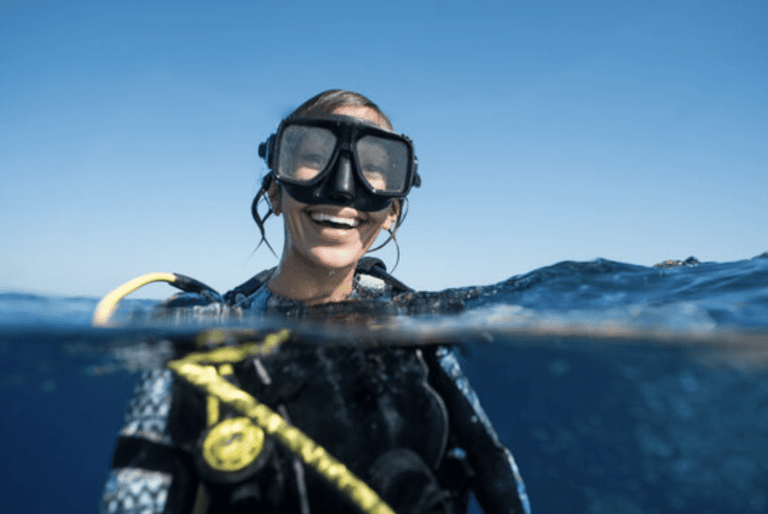 A woman wearing a wetsuit and scuba diving gear smiles above the surface of clear blue water, with goggles on and a bright sky in the background.