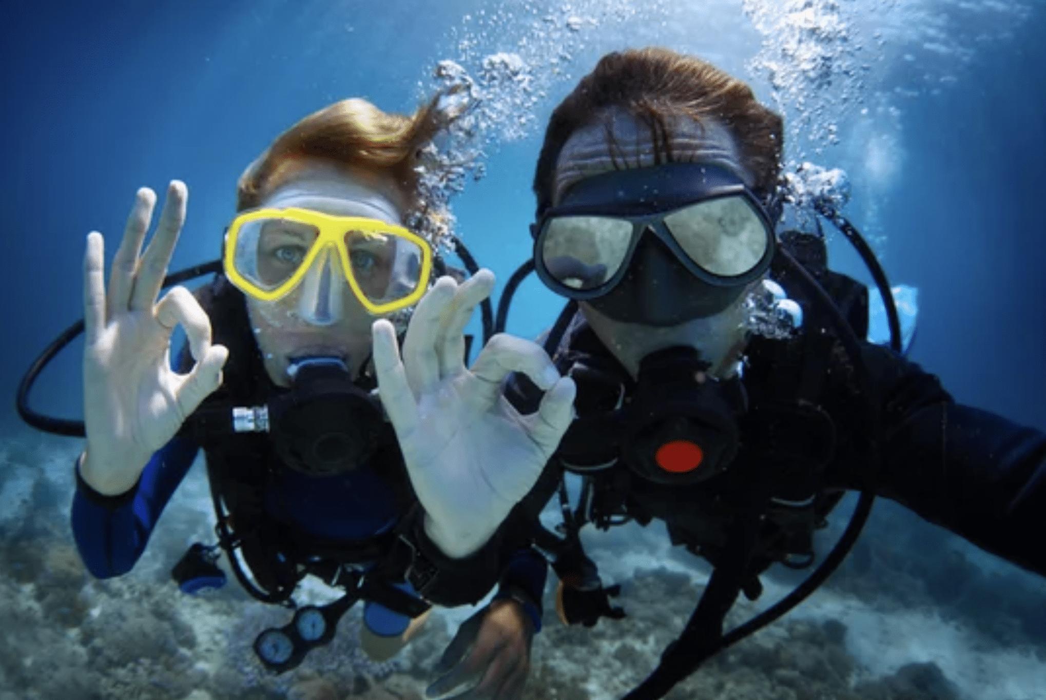 Two scuba divers underwater wearing masks and wetsuits make “OK” hand signals, with bubbles rising around them and coral visible beneath.