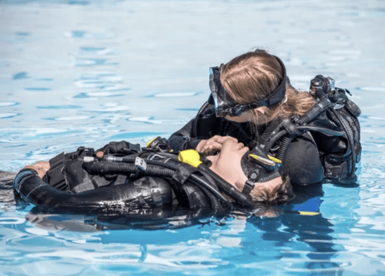 A scuba diver performs a rescue manoeuvre on another diver in a swimming pool, both wearing wetsuits and full scuba gear. The rescuer supports the other diver, who appears unconscious and is floating on their back.