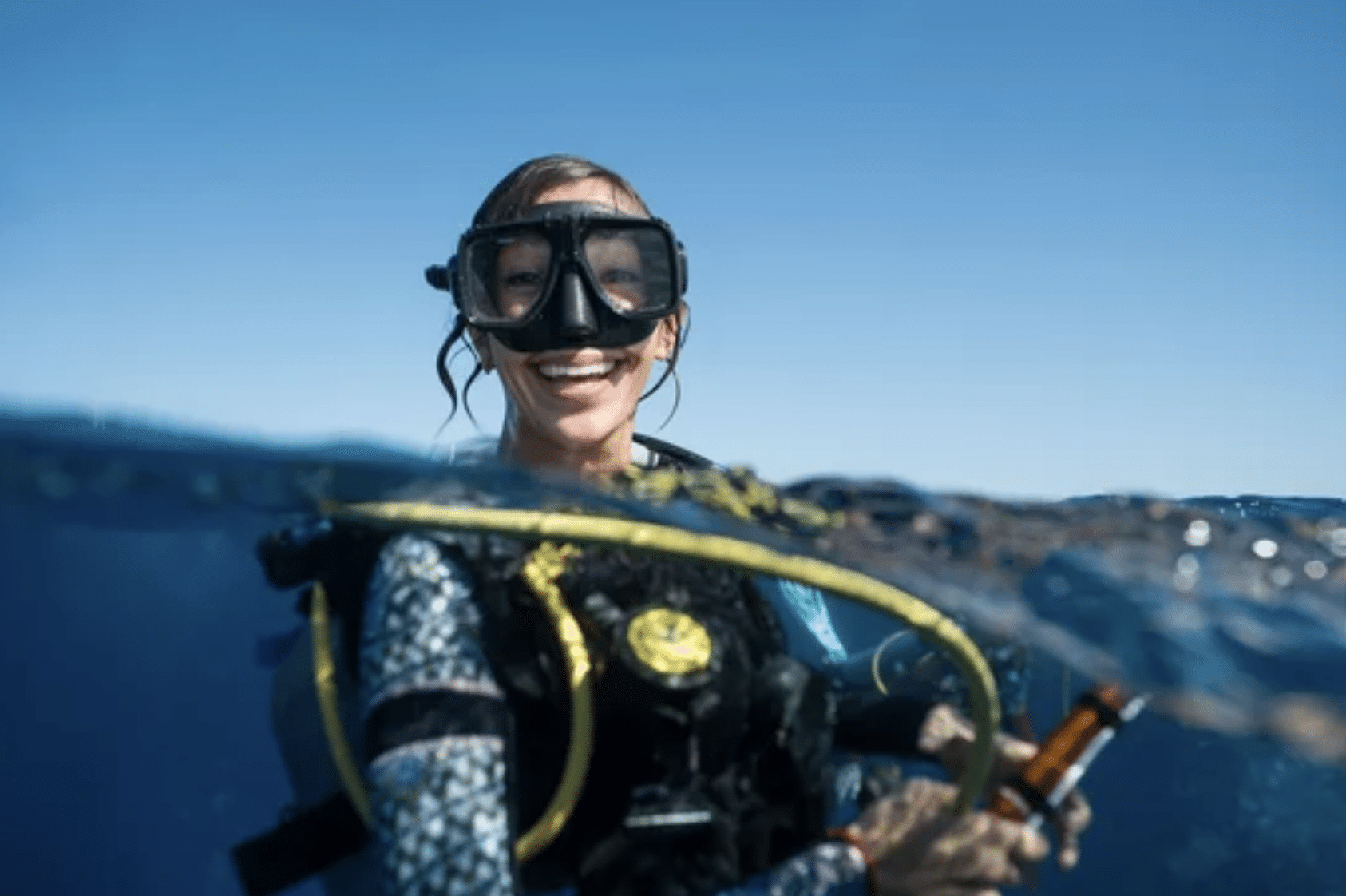 A smiling scuba diver in a wetsuit and mask is partially submerged in blue ocean water, holding equipment, with a clear sky in the background.