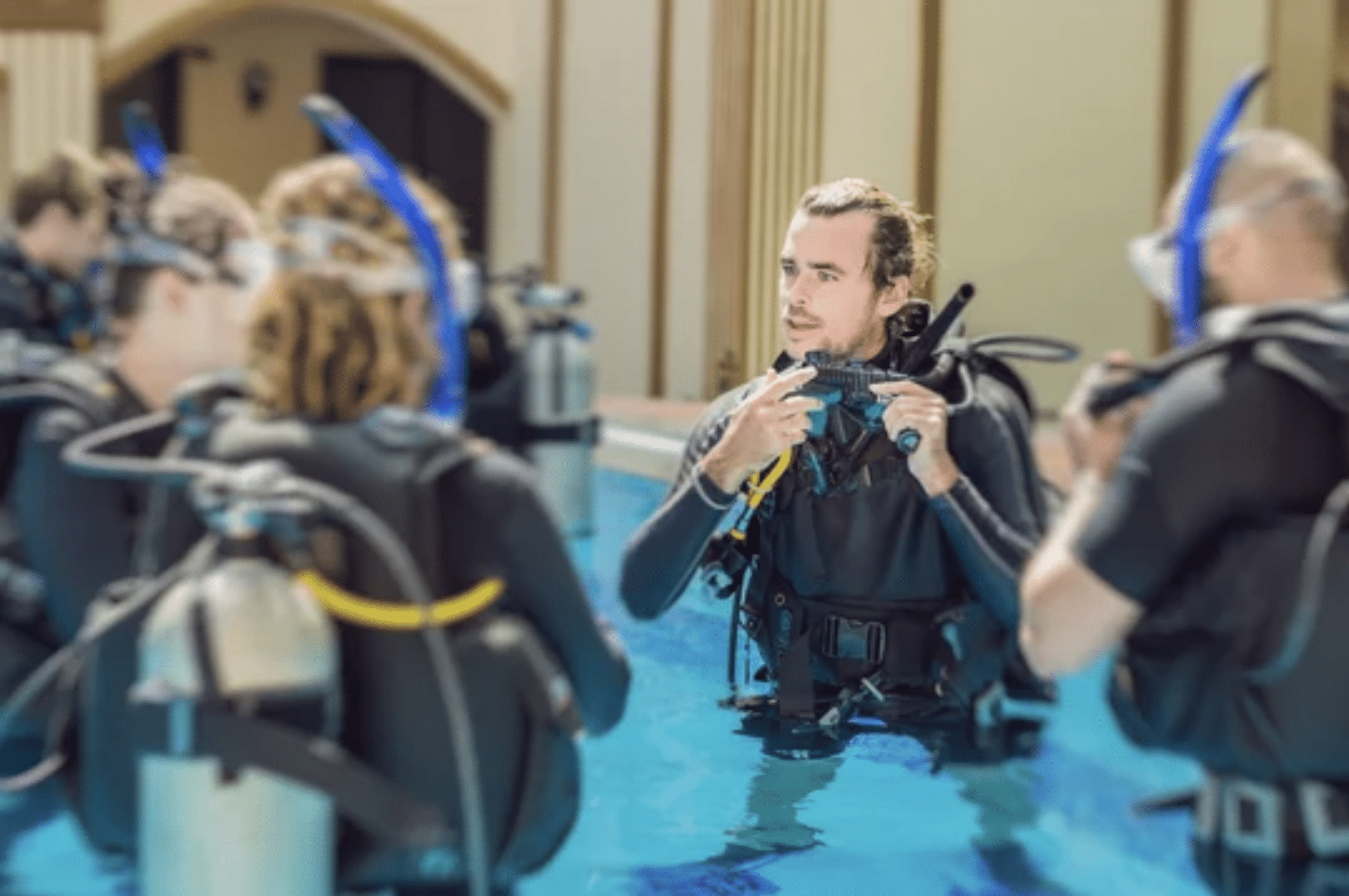 A scuba diving instructor stands in a swimming pool holding a mask and regulator, explaining equipment to three students in wetsuits and scuba gear, preparing for a diving lesson in an outdoor setting.