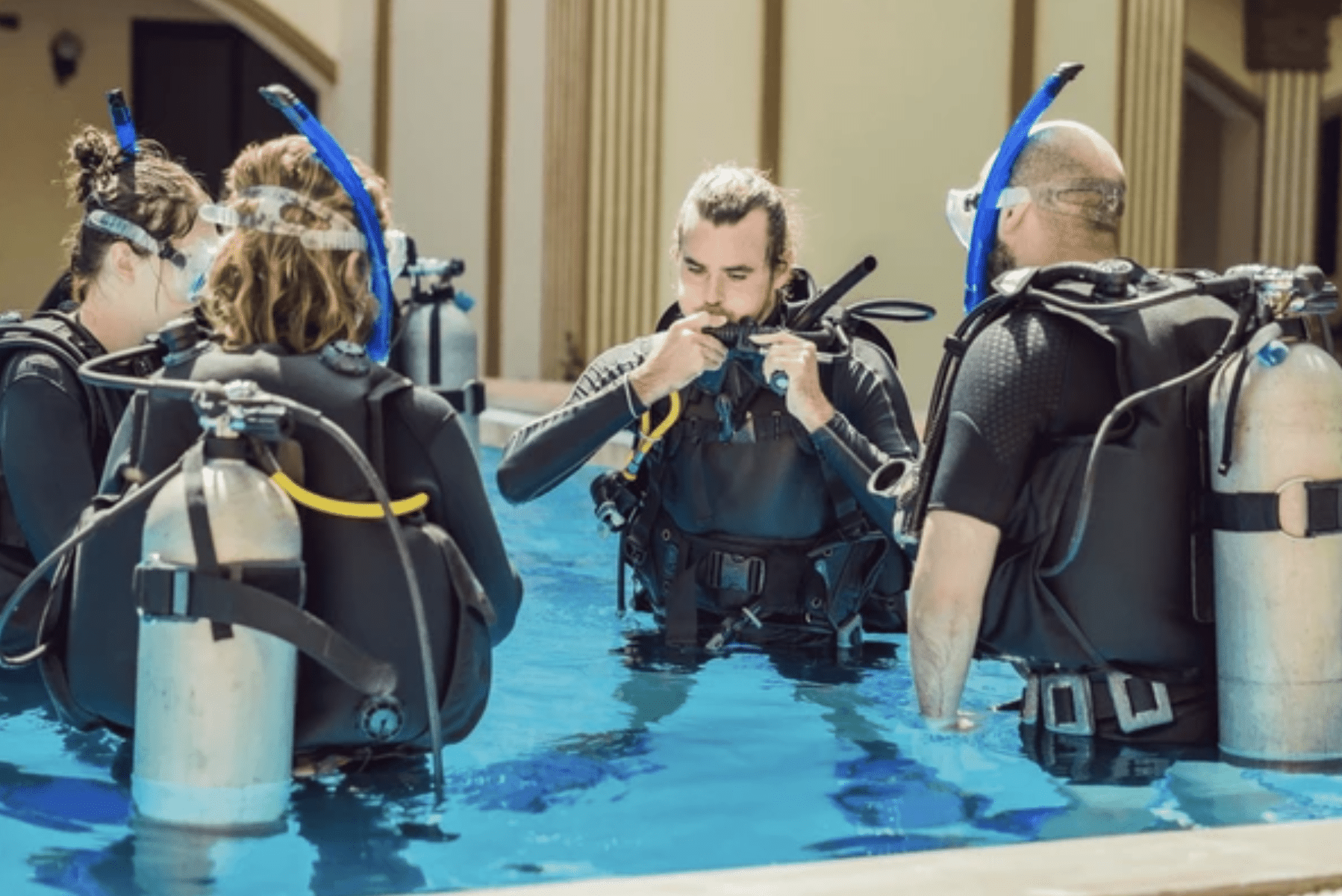 A group of four people in scuba gear stand in a pool, listening to an instructor demonstrating equipment use during a scuba diving lesson.