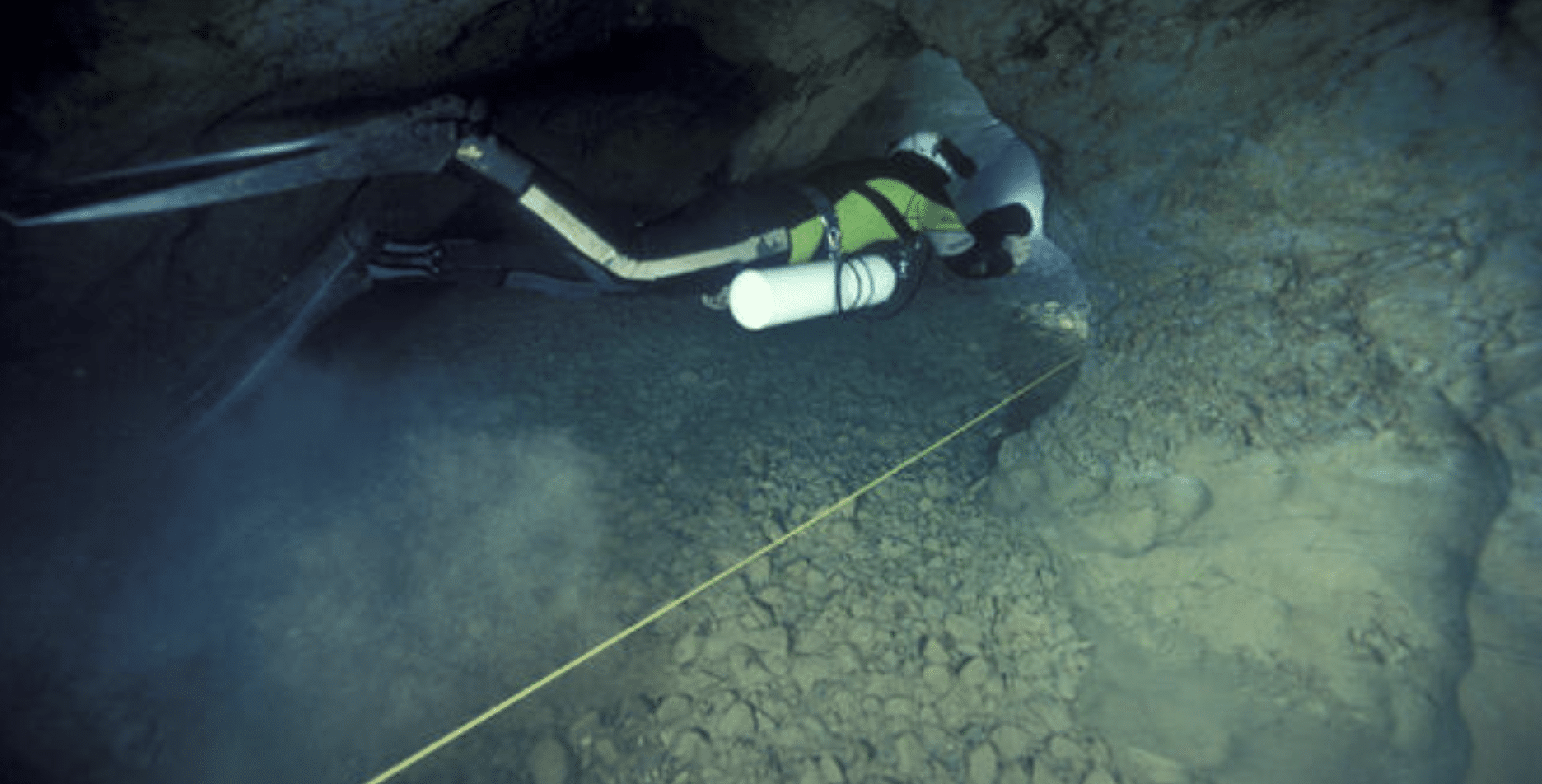 Screenshot 2026-05-23 at 232003 - N9BO℠ | Global Underwater Services Ltd A scuba diver with oxygen cylinders explores a narrow underwater cave, following a guideline stretched along the rocky cave floor.