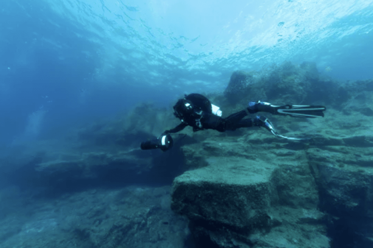 Scuba diver in a black wetsuit with an underwater camera rig, exploring a rocky blue ocean environment.