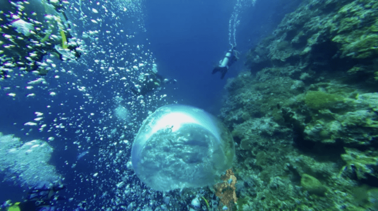 Two scuba divers swim near a coral-covered underwater cliff, surrounded by bubbles. A large, clear air bubble floats in the centre of the deep blue water, reflecting light and the rocky landscape below.