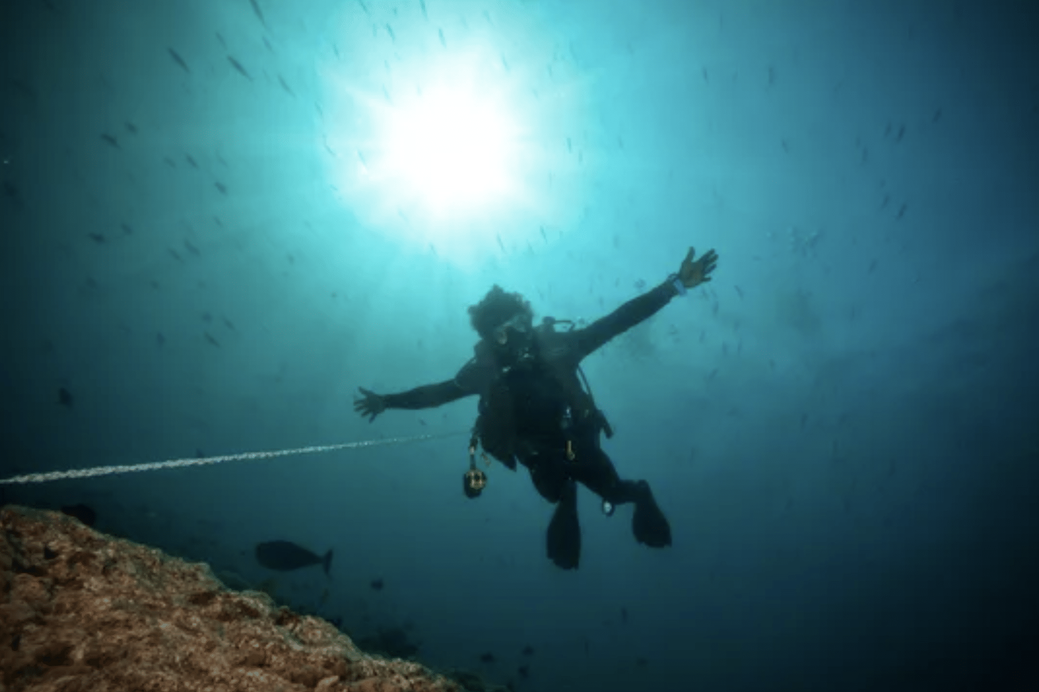 A scuba diver underwater stretches their arms wide, attached to a safety line near a rocky seabed, with sunlight streaming down from the surface above.