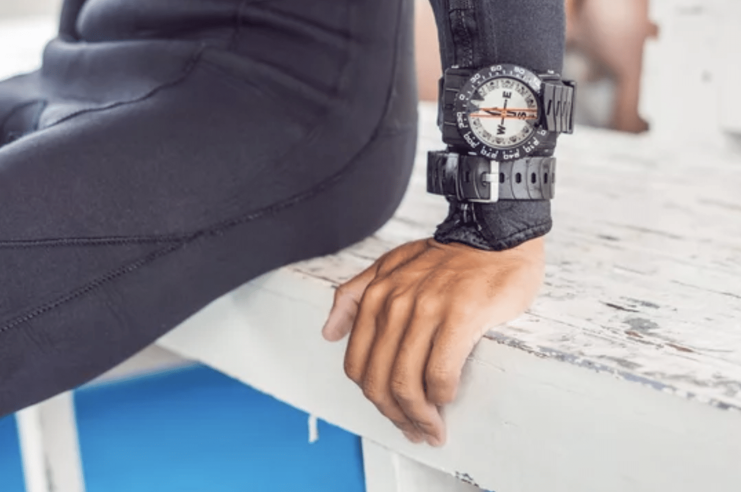 Screenshot 2026-05-27 at 191330 - N9BO℠ | Global Underwater Services Ltd A person in a black wetsuit sits on a white, weathered bench with their hand resting on the surface. They are wearing a large black wristwatch over the sleeve of the wetsuit.