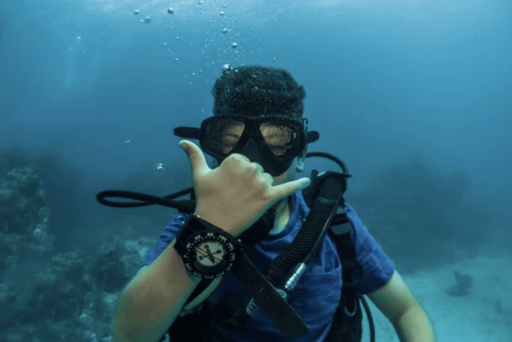 Screenshot 2026-05-27 at 191405 - N9BO℠ | Global Underwater Services Ltd A scuba diver underwater in clear blue water makes a shaka hand sign, wearing a diving mask, snorkel, and a wrist compass, with bubbles rising above and coral visible in the background.