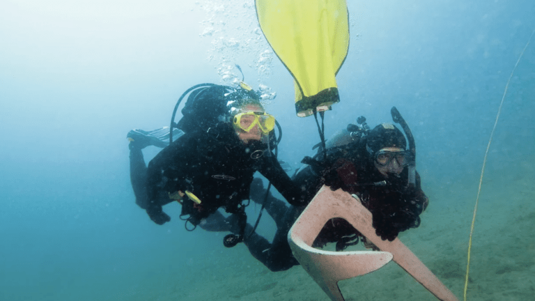 Two divers in full kit explore underwater next to a yellow buoy and a large metal object.
