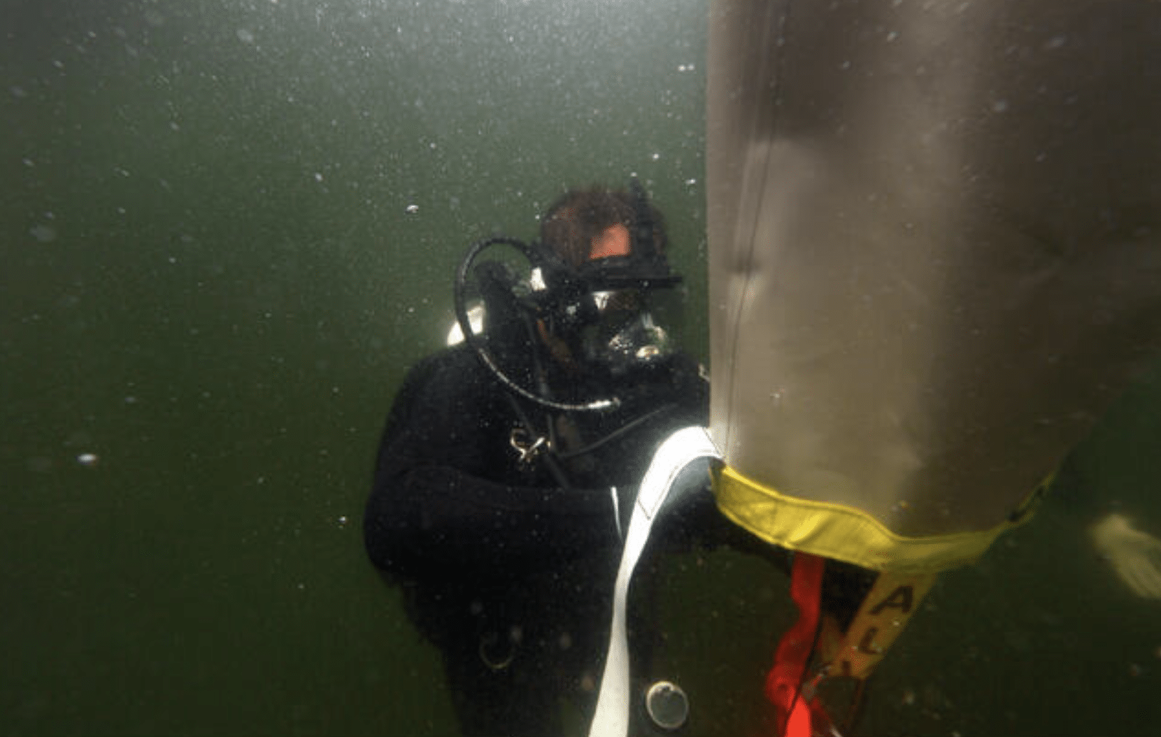 Screenshot 2026-05-27 at 225758 - N9BO℠ | Global Underwater Services Ltd A scuba diver underwater in murky greenish water, wearing full diving kit and holding onto a large, partially inflated lift bag with yellow and white straps.