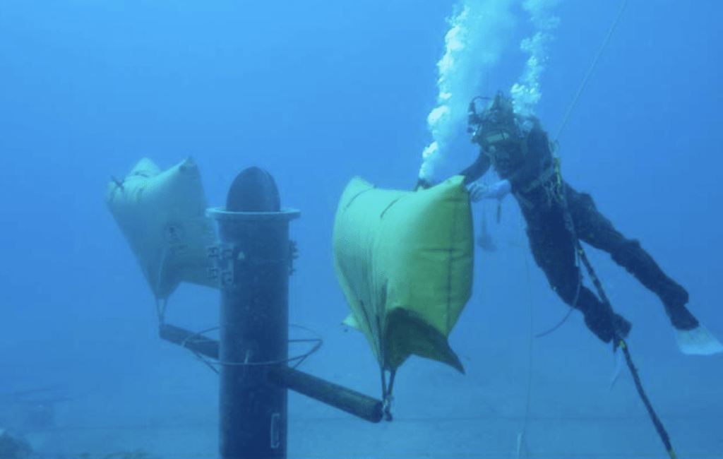 Screenshot 2026-05-27 at 230001 - N9BO℠ | Global Underwater Services Ltd A diver underwater attaches large yellow lifting bags to a submerged metal structure while bubbles rise from the diver’s equipment. The scene takes place in clear blue water, suggesting an underwater construction or salvage operation.