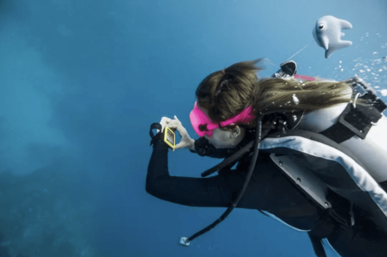 A scuba diver wearing a pink mask and black wetsuit checks a diving computer underwater, with a small, toy-like whale attached to her kit, surrounded by blue ocean water.