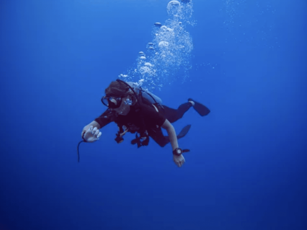 Screenshot 2026-05-28 at 220753 - N9BO℠ | Global Underwater Services Ltd A scuba diver in full kit swims underwater, surrounded by clear blue water and bubbles rising above them. The diver is looking at a device on their wrist and holding a lead in one hand.