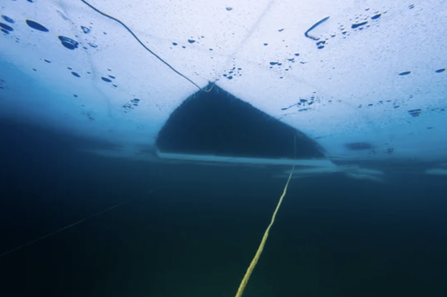 Underwater view showing a large triangular hole in the ice above, with a yellow rope leading up to the opening. Blue tones dominate the scene, suggesting cold temperatures and icy water.