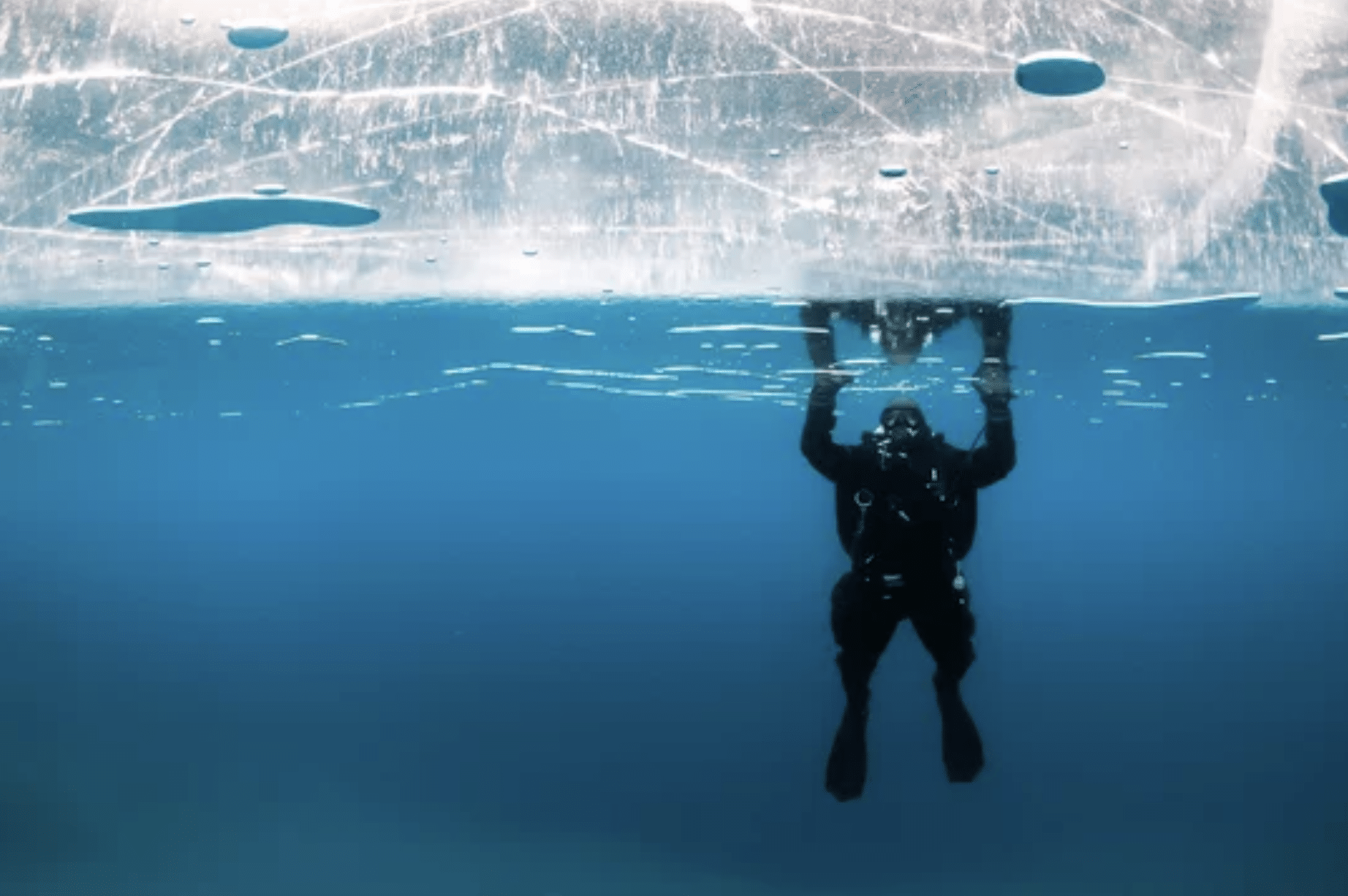 A scuba diver in dark kit is underwater, holding onto the underside of thick, cracked ice, with clear blue water below and light filtering through the ice above.