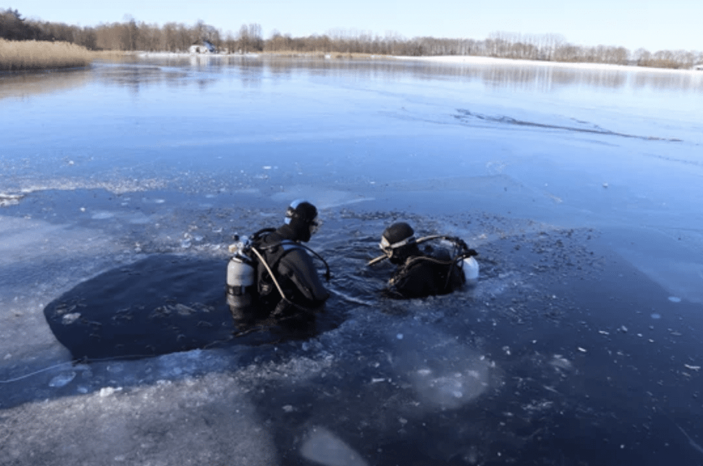Two scuba divers in wetsuits are partially submerged in a hole cut into the surface of a frozen lake, surrounded by ice and snow, with trees and reeds visible in the background on a clear day.