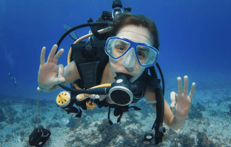 A scuba diver underwater wears a mask and breathing apparatus while making the “OK” hand signal with both hands. The diver is surrounded by clear blue water and a rocky sea floor is visible below.