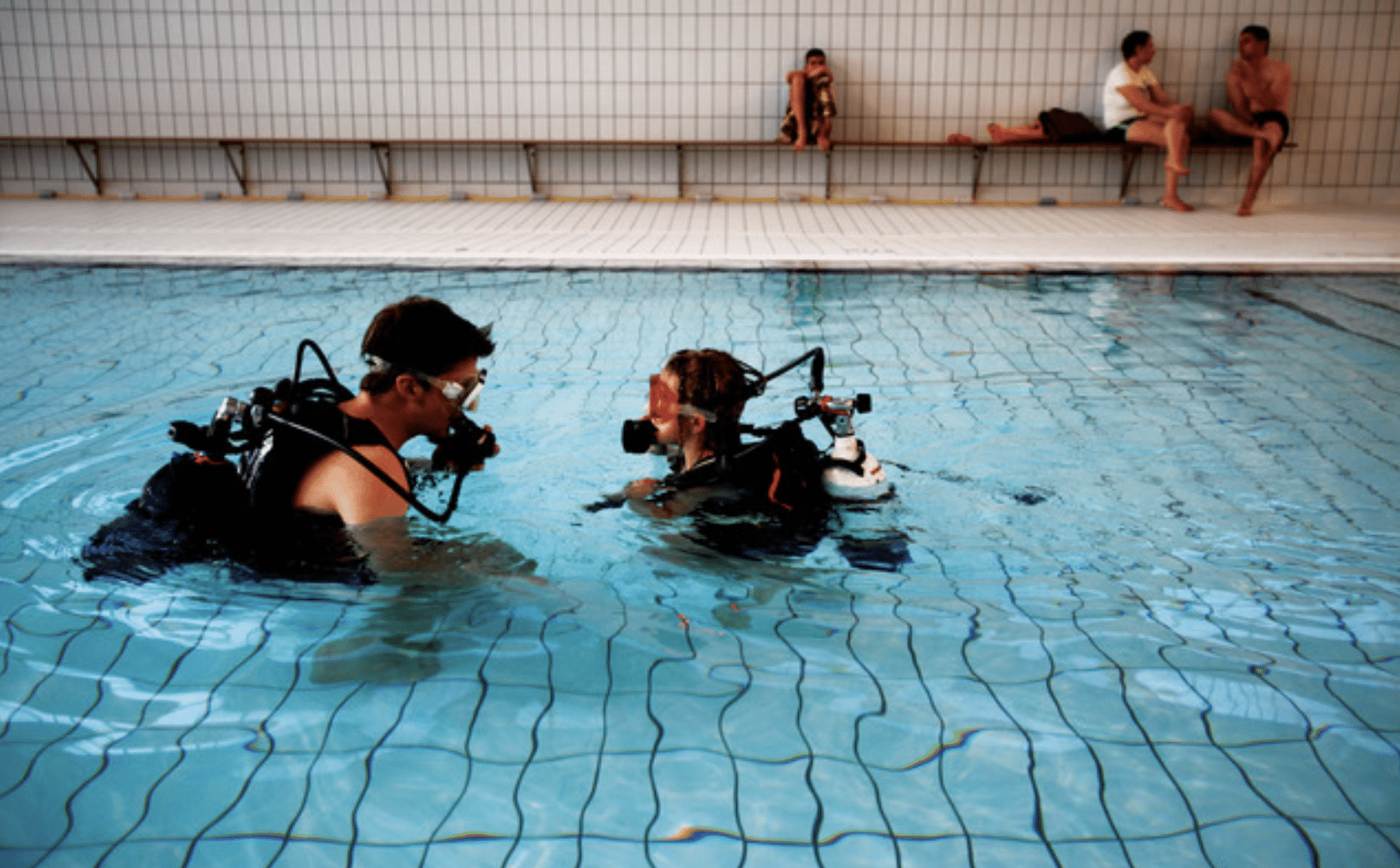 Screenshot 2026-06-02 at 065909 - N9BO℠ | Global Underwater Services Ltd Two people wearing scuba gear practise in an indoor swimming pool, facing each other. Three others sit on benches by the poolside in the background, observing the activity.