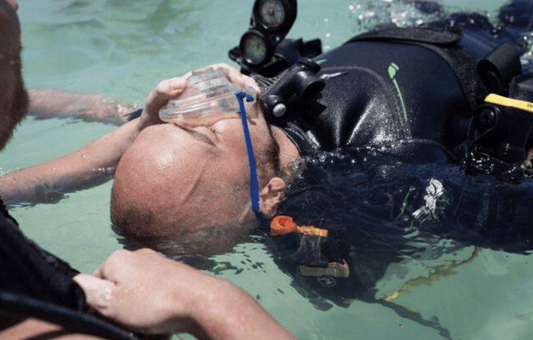 A scuba diver in black gear floats on their back in shallow water while another person holds an oxygen mask to the diver’s face, suggesting a rescue or medical emergency.