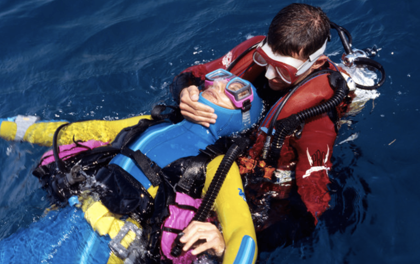 A scuba diver in a red wetsuit supports and rescues another diver wearing a blue and yellow wetsuit, floating on the surface of the water. The rescuer holds the injured diver's head and mask above water.