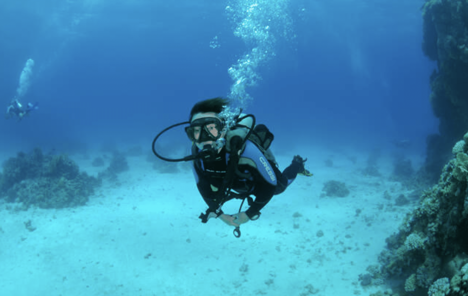 Screenshot 2026-06-03 at 161010 - N9BO℠ | Global Underwater Services Ltd A scuba diver swims underwater near a coral reef, surrounded by clear blue water. Bubbles rise from the diver’s breathing apparatus, and another diver can be seen in the distance.