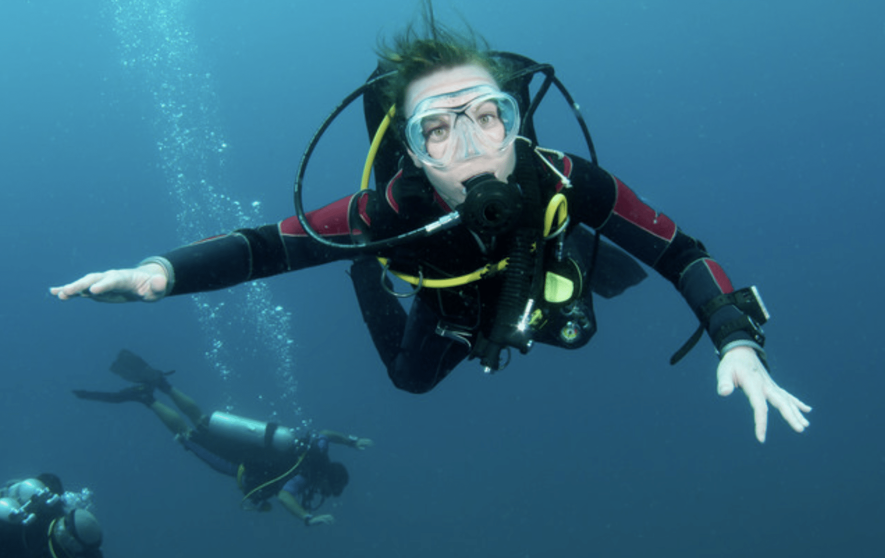 Screenshot 2026-06-03 at 161037 - N9BO℠ | Global Underwater Services Ltd A scuba diver in a wetsuit and mask swims underwater, facing the camera with arms outstretched. Two more divers are visible in the background, surrounded by blue water and rising bubbles.