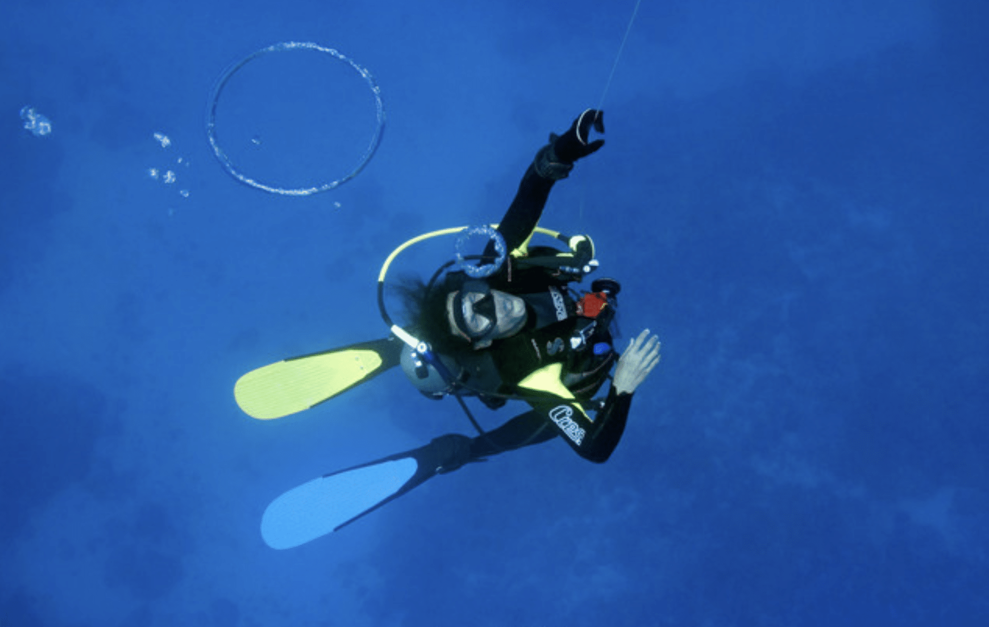 A scuba diver wearing yellow and blue flippers floats underwater, reaching upwards with one hand, surrounded by bubbles and deep blue water.