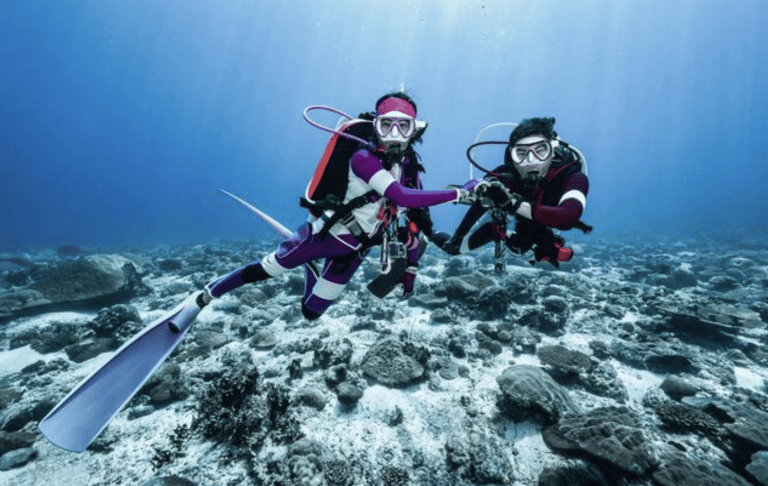 Two scuba divers in wetsuits are underwater above a coral reef, holding hands and looking at the camera. Sunlight filters through the clear blue water, illuminating the rocky seabed below them.