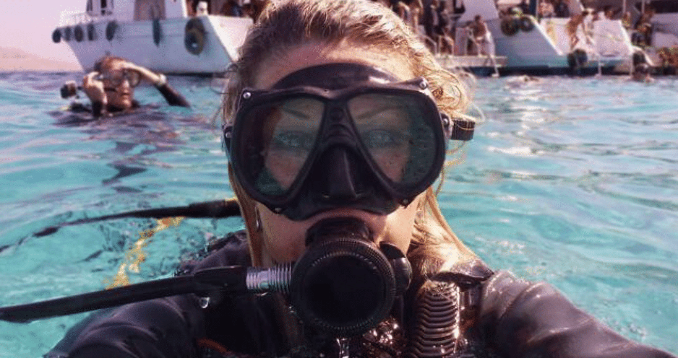 A person wearing scuba diving kit, including a mask and regulator, takes a selfie whilst floating in clear blue water near boats with other divers visible in the background.