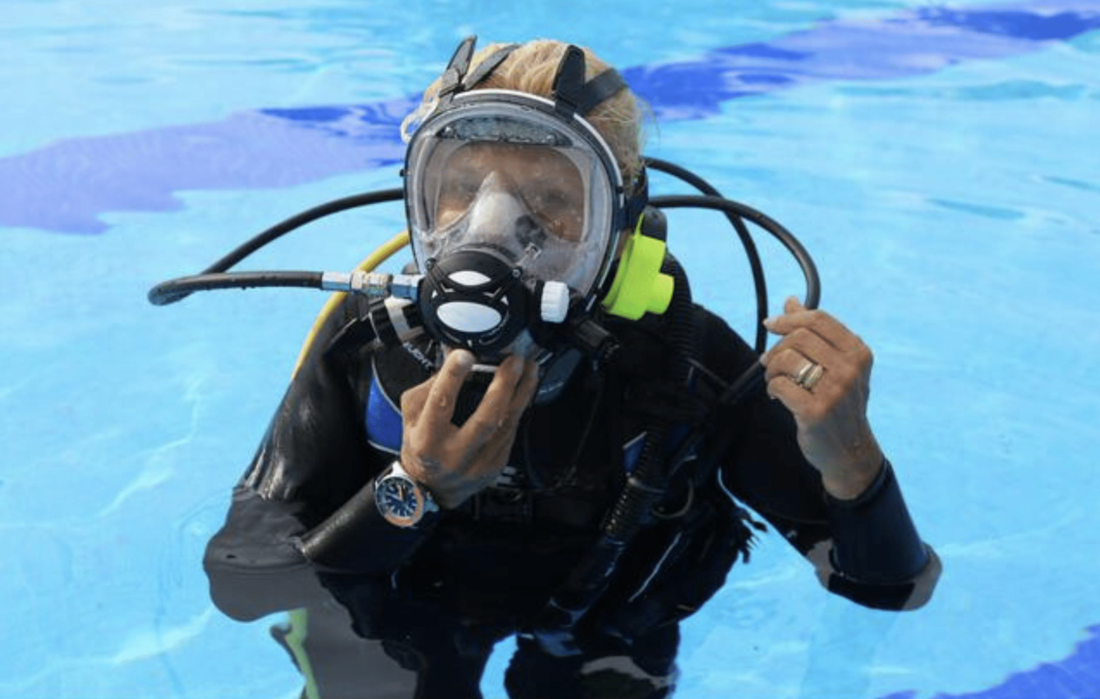 A person in full scuba diving gear, including a mask and air tank, is partially submerged in a clear blue swimming pool, adjusting their mask with one hand.